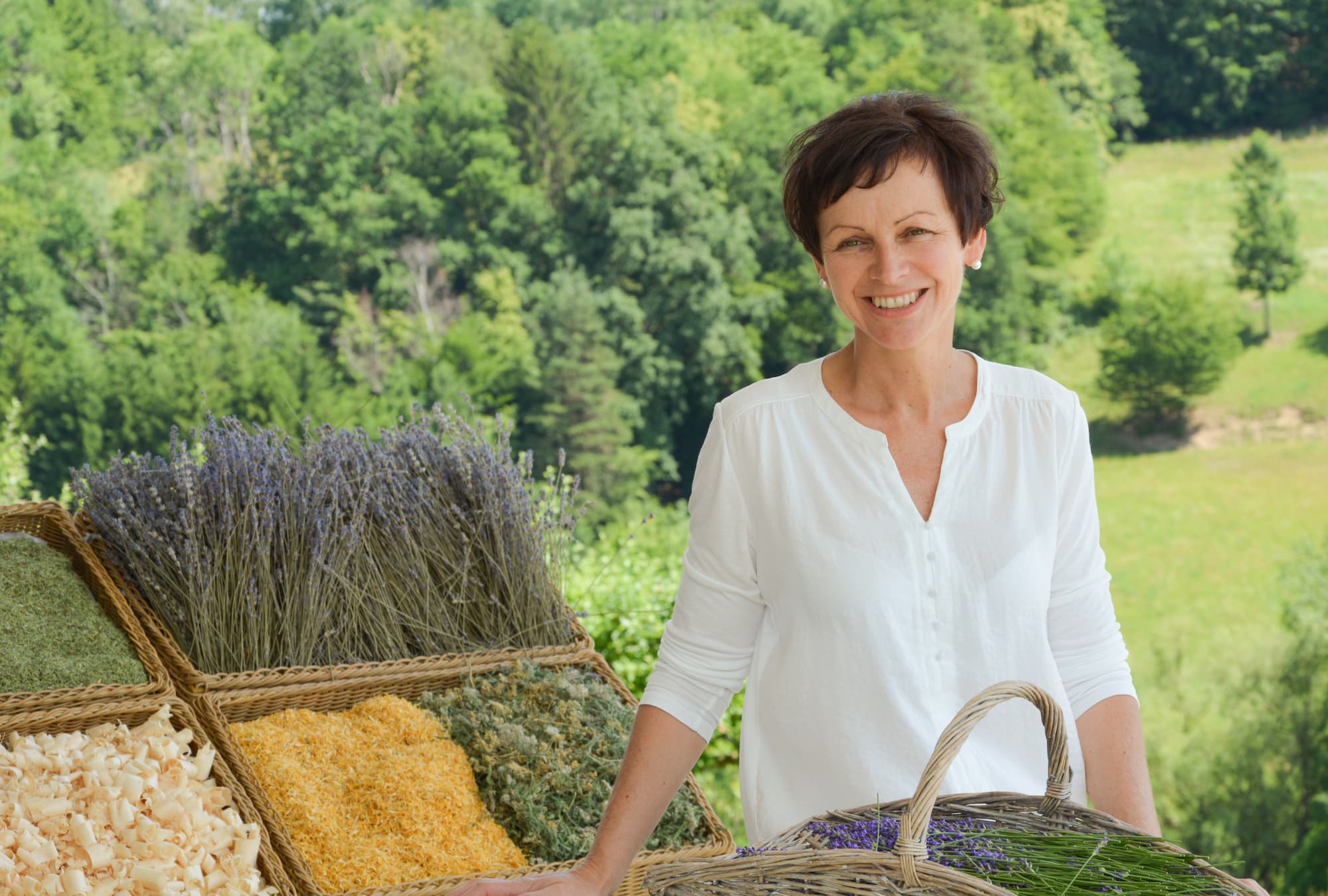 Eine Frau Präsentiert frische Kreuter in Körben, im Hintergrund ein Waldstück mit Wiese